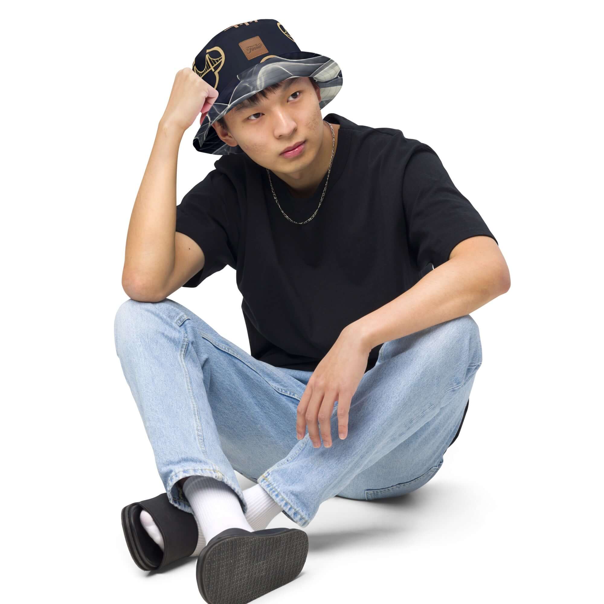 Young man wearing a reversible bucket hat streetwear, paired with casual jeans and a black t-shirt, sitting down confidently.